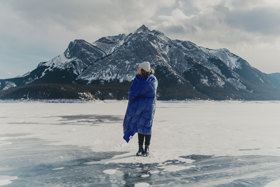 Person wrapped in a Nakie puffy blanket standing on a frozen lake with snow-covered mountains in the background