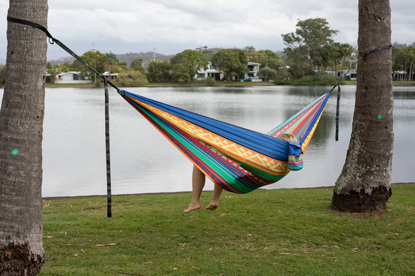 Colorful striped recycled hammock with black straps hung between two trees overlooking water and houses.