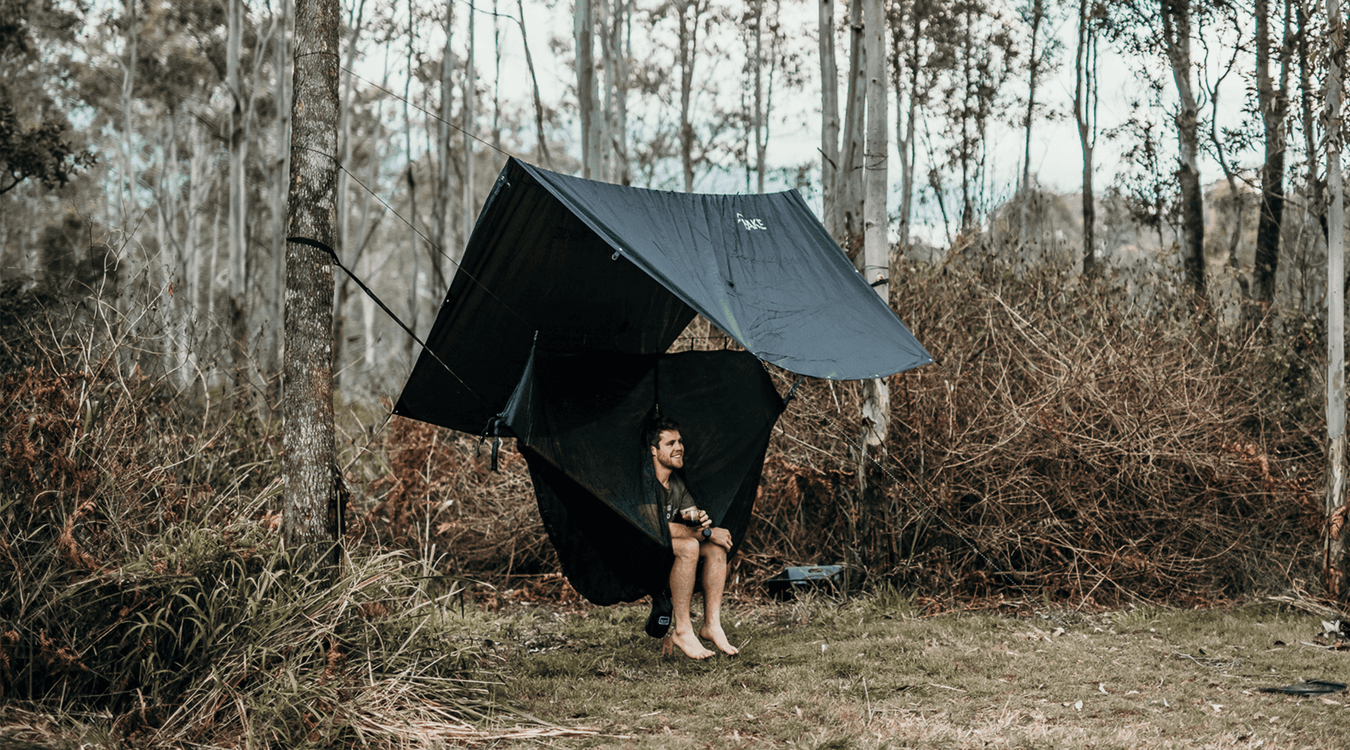 Person snug in nakie Hammock protected by a Nakie Rain tarp in the woods.