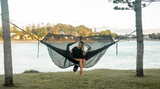 Woman in a Nakie hammock with a bugnet relaxing by the water. Nakie - Australia