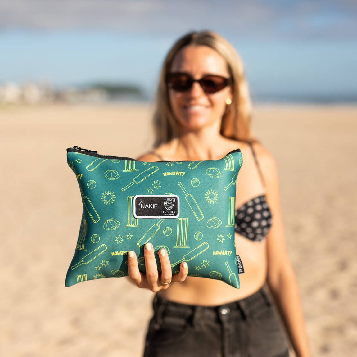 Teal recycled sand-free beach towel with cricket-themed gold pattern, held up on beach by woman wearing sunglasses