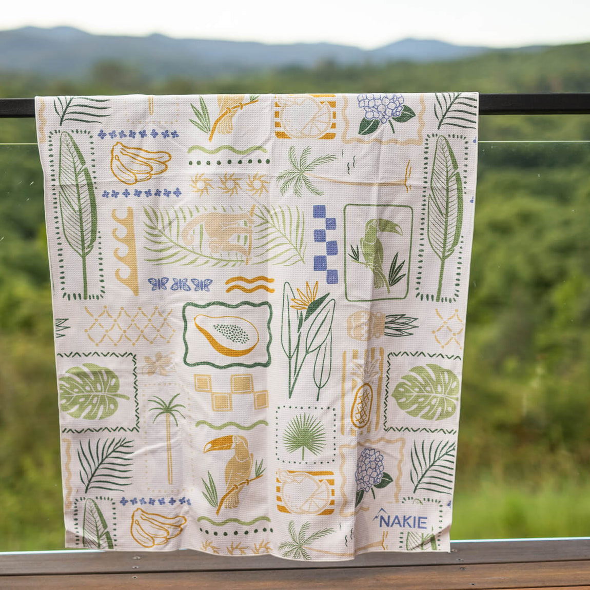 Cream beach towel with tropical botanical print featuring toucans, papayas, palm leaves, and geometric patterns in green