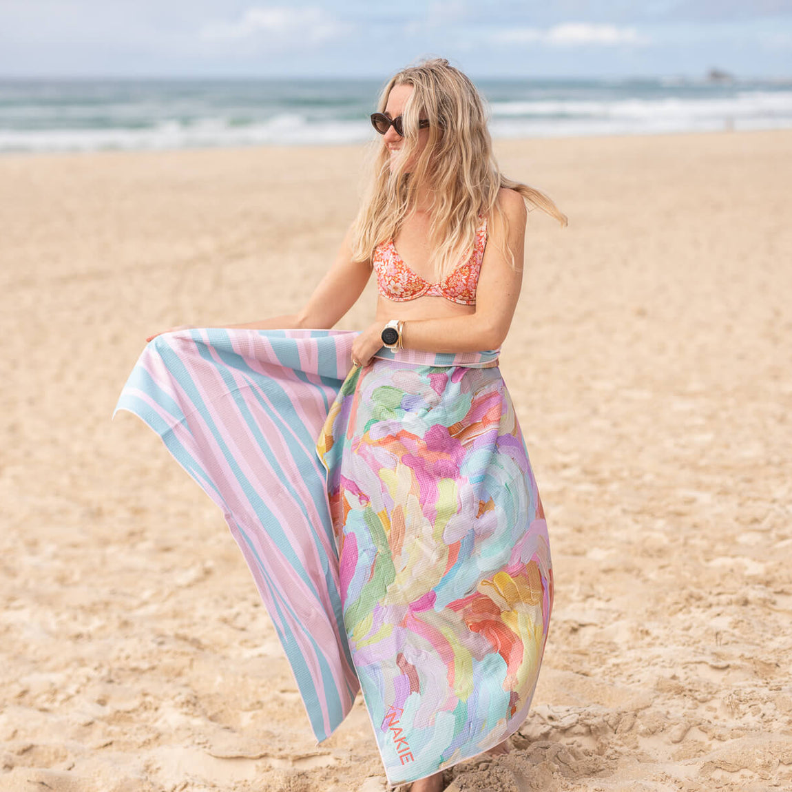 Recycled sand-free beach towel with colorful floral print, shown held on beach with striped reverse side visible.