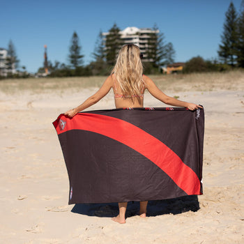 Black and red beach towel with diagonal stripe design, displayed on sand by person facing away from camera