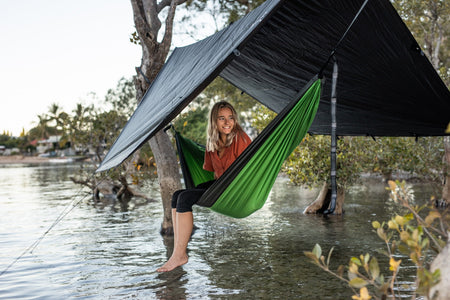 Woman sheltering under a Nakie tarp made from recycled plastic bottles, enjoying a dry outdoor experience.