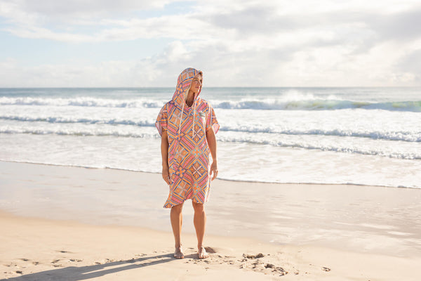 Coral geometric patterned hooded towel with short sleeves worn on beach, sand-free fabric visible.