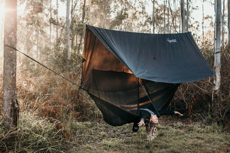 Woman lying comfortably in a Nakie hammock, covered by our ultralight no knot tarp, enjoying the sunrise and the cozy embrace of sustainable outdoor gear.