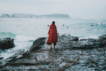 Image of a man wrapped in Nakie's Earth Red Sustainable Down Puffy Blanket, watching other people swim, enjoying warmth and sustainability by the water. preview #6