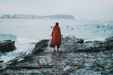 Image of a man wrapped in Nakie's Earth Red Sustainable Down Puffy Blanket, watching other people swim, enjoying warmth and sustainability by the water.