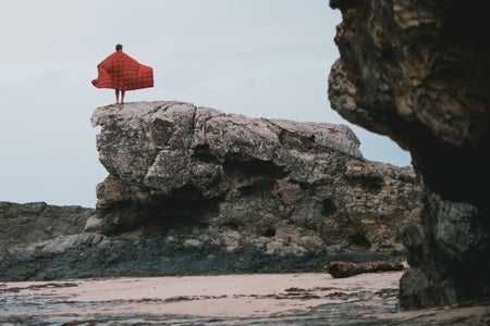 A man draping Nakie's Earth Red Sustainable Down Puffy Blanket over her back while admiring the beach view.