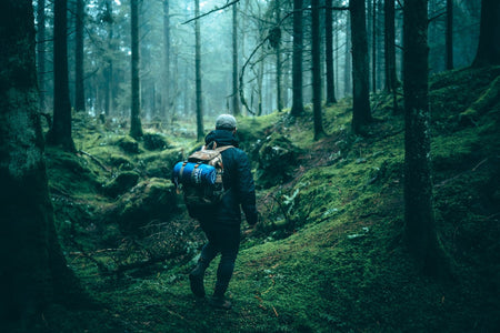 Image of a man walking in a campsite with Nakie's Deep Ocean Blue Sustainable Down Puffy Blanket attached to his backpack, ready for outdoor adventures with sustainable warmth.