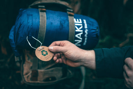 Unopened Nakie's Deep Ocean Blue Sustainable Down Puffy Blanket attached to the front of a bag, with a person's hand holding the product tag displaying the recycled symbol, highlighting its eco-friendly design.