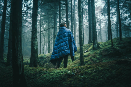 Image of a man wrapped in Nakie's Deep Ocean Blue Sustainable Down Puffy Blanket while standing in a campsite, enjoying warmth and sustainability during outdoor adventures.