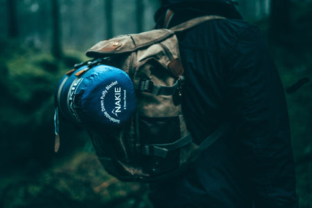 Unopened Nakie's Deep Ocean Blue Sustainable Down Puffy Blanket attached to the front of a backpack worn by a man in a campsite, showcasing its portability and eco-friendly design.