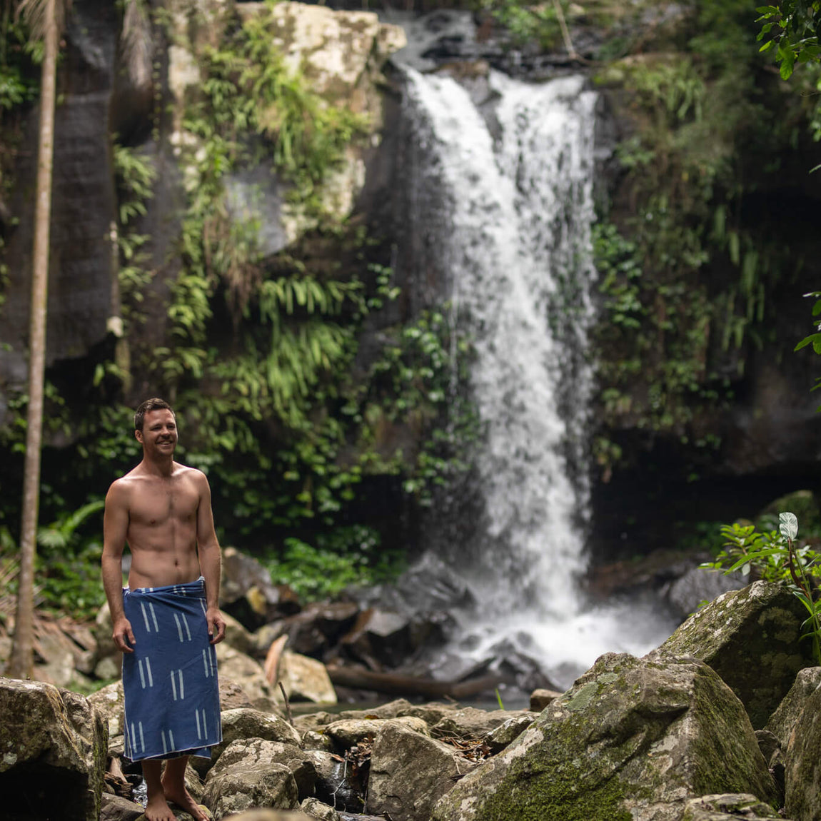 Navy beach towel with white stripe pattern, worn as wrap by man standing on rocks near waterfall