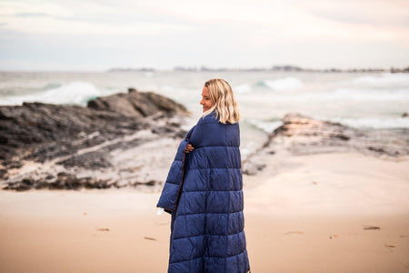 Image of a woman standing on a beach, wrapped in Nakie's Deep Ocean Blue Sustainable Down Puffy Blanket, enjoying warmth and sustainability during her beach outing.