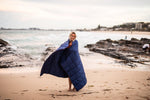 Image of a woman standing on a beach, wrapped in Nakie's Deep Ocean Blue Sustainable Down Puffy Blanket, enjoying warmth and sustainability during her beach outing. preview #11