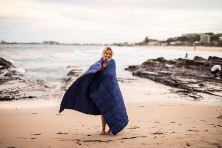 Image of a woman standing on a beach, wrapped in Nakie's Deep Ocean Blue Sustainable Down Puffy Blanket, enjoying warmth and sustainability during her beach outing.