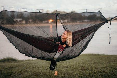 A smiling woman peeking out from inside Nakie's Bug Net while lying in a hammock, enjoying protected relaxation from insects during outdoor adventures.