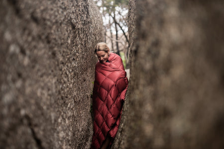 Image of a woman wrapped in Nakie's Earth Red Sustainable Down Puffy Blanket, enjoying warmth and sustainability during outdoor adventures.