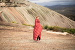 Image of a smiling woman wrapped in Nakie's Earth Red Sustainable Down Puffy Blanket, enjoying warmth and comfort during outdoor adventures. preview #9