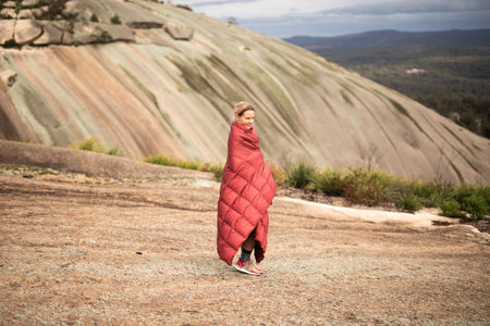 Image of a smiling woman wrapped in Nakie's Earth Red Sustainable Down Puffy Blanket, enjoying warmth and comfort during outdoor adventures.