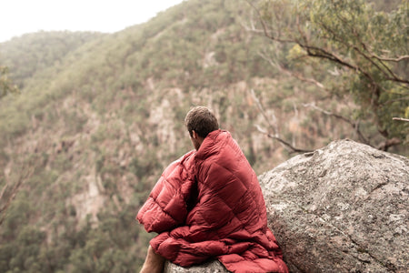 Image of a man wrapped in Nakie's Earth Red Sustainable Down Puffy Blanket, enjoying the mountain view while sitting on a cliff, embracing warmth and sustainability during outdoor exploration.