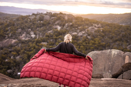 Image of a woman holding Nakie's Earth Red Sustainable Down Puffy Blanket behind her back, showcasing its vibrant color and sustainable design.