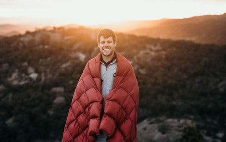 Image of a man wrapped in Nakie's Earth Red Sustainable Down Puffy Blanket, enjoying warmth and sustainability during outdoor adventures.