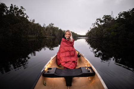 Image of a woman wrapped in Nakie's Earth Red Sustainable Down Puffy Blanket, sitting on a boat and enjoying warmth and sustainability during her maritime adventure.