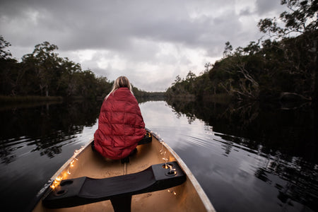 Image of a woman wrapped in Nakie's Earth Red Sustainable Down Puffy Blanket, sitting on a boat and enjoying warmth and sustainability during her maritime adventure.