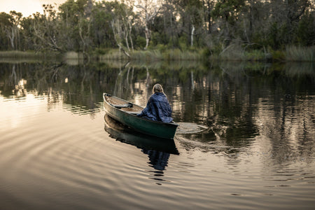 Image of a woman wrapped in Nakie's Deep Ocean Blue Sustainable Down Puffy Blanket while on a boat, enjoying warmth and sustainability during her maritime adventure.