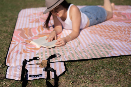 Woman enjoying a book at a Nakie picnic, surrounded by nature and relaxation, embodying the brand's commitment to outdoor leisure and sustainability.