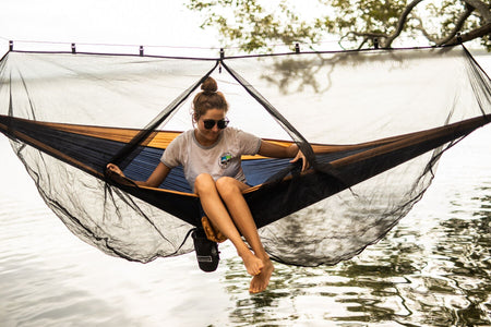 A woman sitting in a hammock in a mangrove forest, with Nakie's Bug Net opened, enjoying the natural surroundings while being prepared for insect protection.